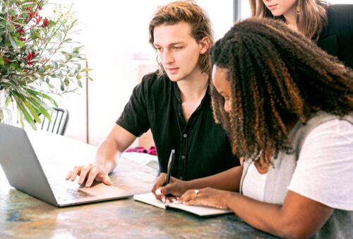 Deux collègues en réunion, l'une prend des notes à la main pendant que l'autre travaille sur un portable